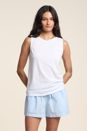 A woman stands against a plain background with her hands behind her back, wearing Velvet by Graham & Spencer’s TAURUS TANK TOP—a white sleeveless crew neck crafted from textured cotton slub—and light blue shorts, creating an essential look for effortless summer style.