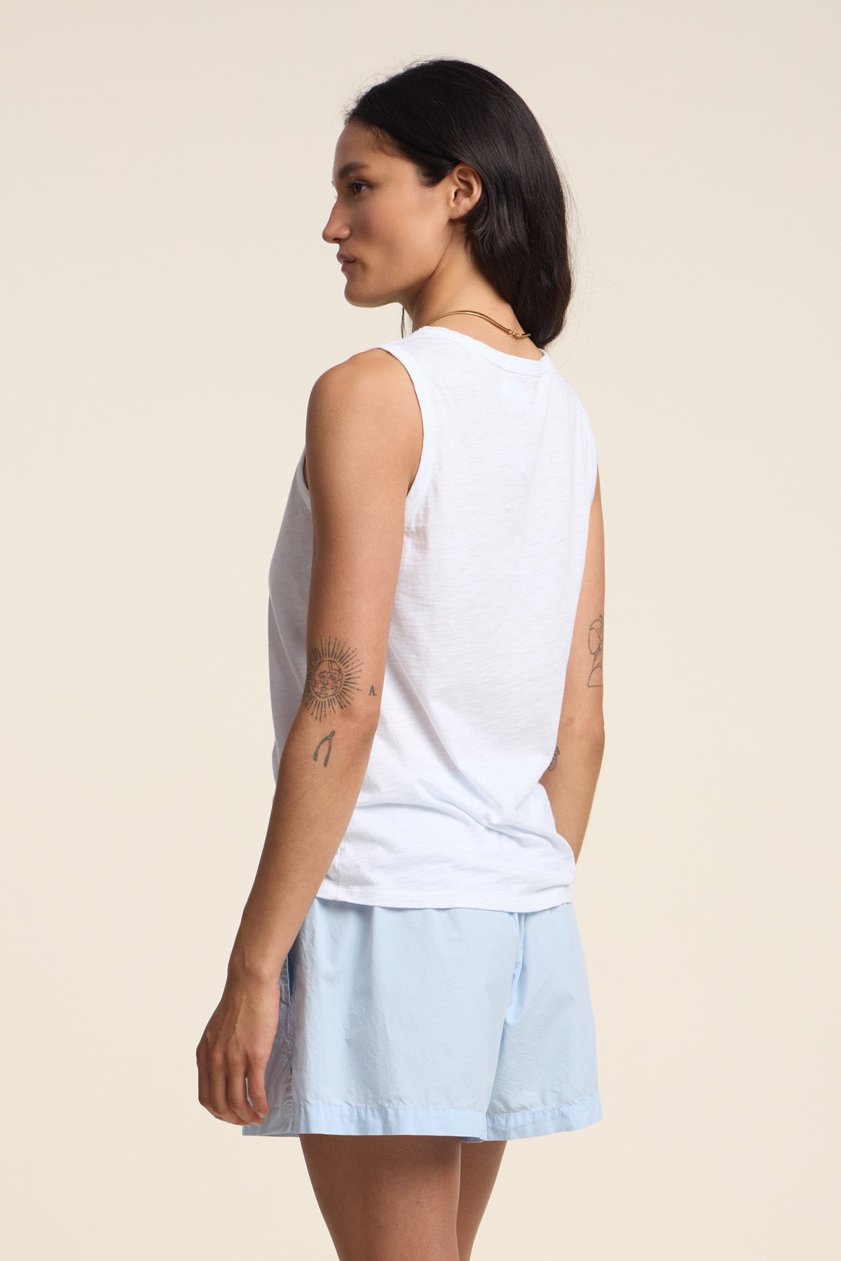 A woman stands sideways against a plain light background, wearing the TAURUS TANK TOP by Velvet by Graham & Spencer—a sleeveless crew neck white top crafted from textured cotton slub—paired with light blue shorts and revealing tattoos on her arm.