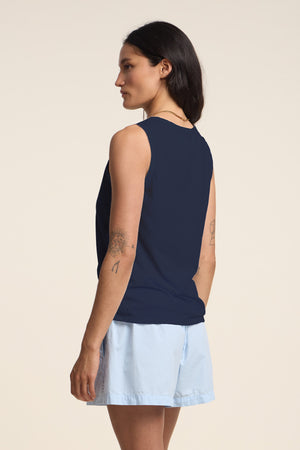 Woman standing sideways in the TAURUS TANK TOP by Velvet by Graham & Spencer—a sleeveless navy textured cotton slub top with a crew neck—paired with light blue shorts. Tattoos visible on her upper arm and forearm, set against a plain light background.