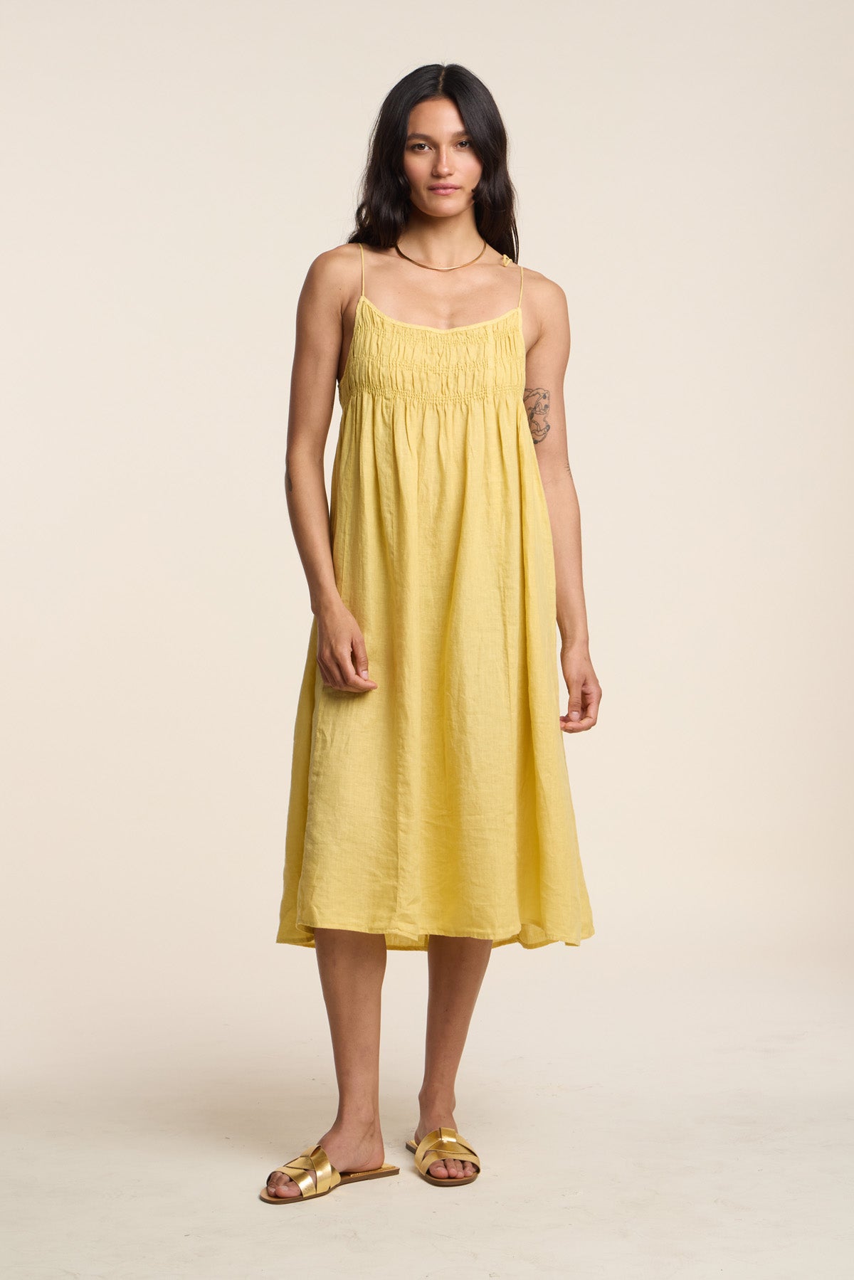A woman stands facing forward wearing the TERESA LINEN DRESS by Velvet by Graham & Spencer—a sleeveless, loose-fitting yellow linen A-line midi dress—paired with gold sandals against a plain beige background.-41527117775041