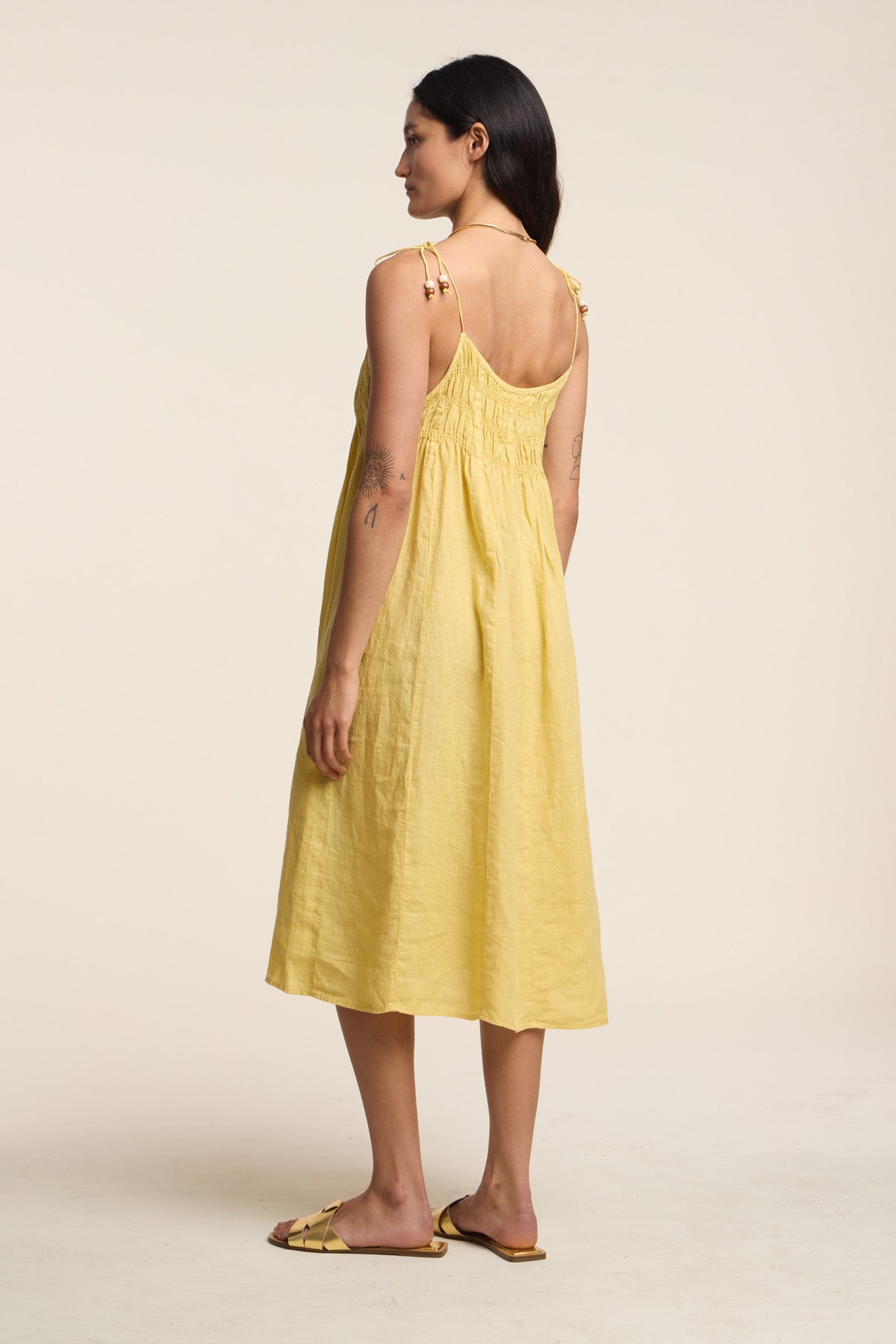 A woman stands sideways on a plain background, wearing Velvet by Graham & Spencer's TERESA LINEN DRESS—a light yellow smocked bust, A-line silhouette dress with shoulder ties—paired with flat sandals.-41527118069953