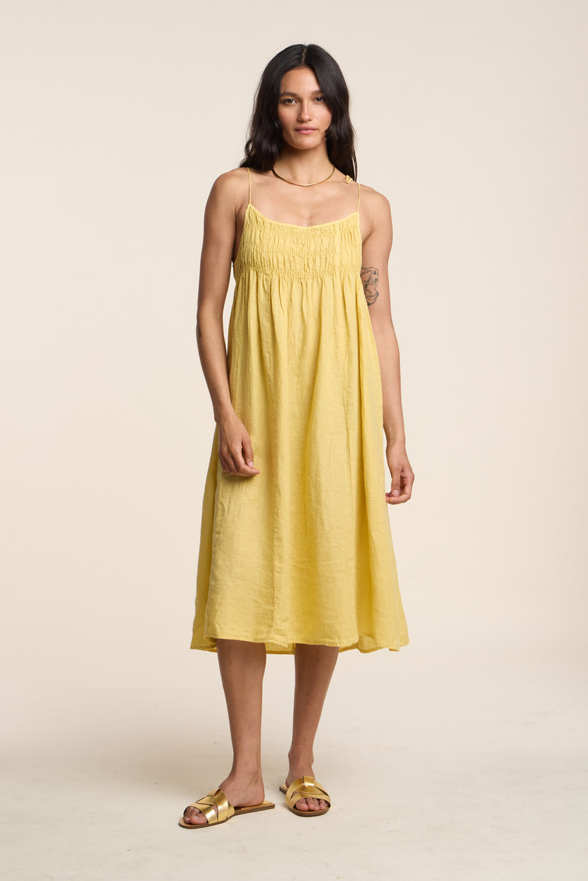 A woman stands facing forward wearing the TERESA LINEN DRESS by Velvet by Graham & Spencer—a sleeveless, loose-fitting yellow linen A-line midi dress—paired with gold sandals against a plain beige background.