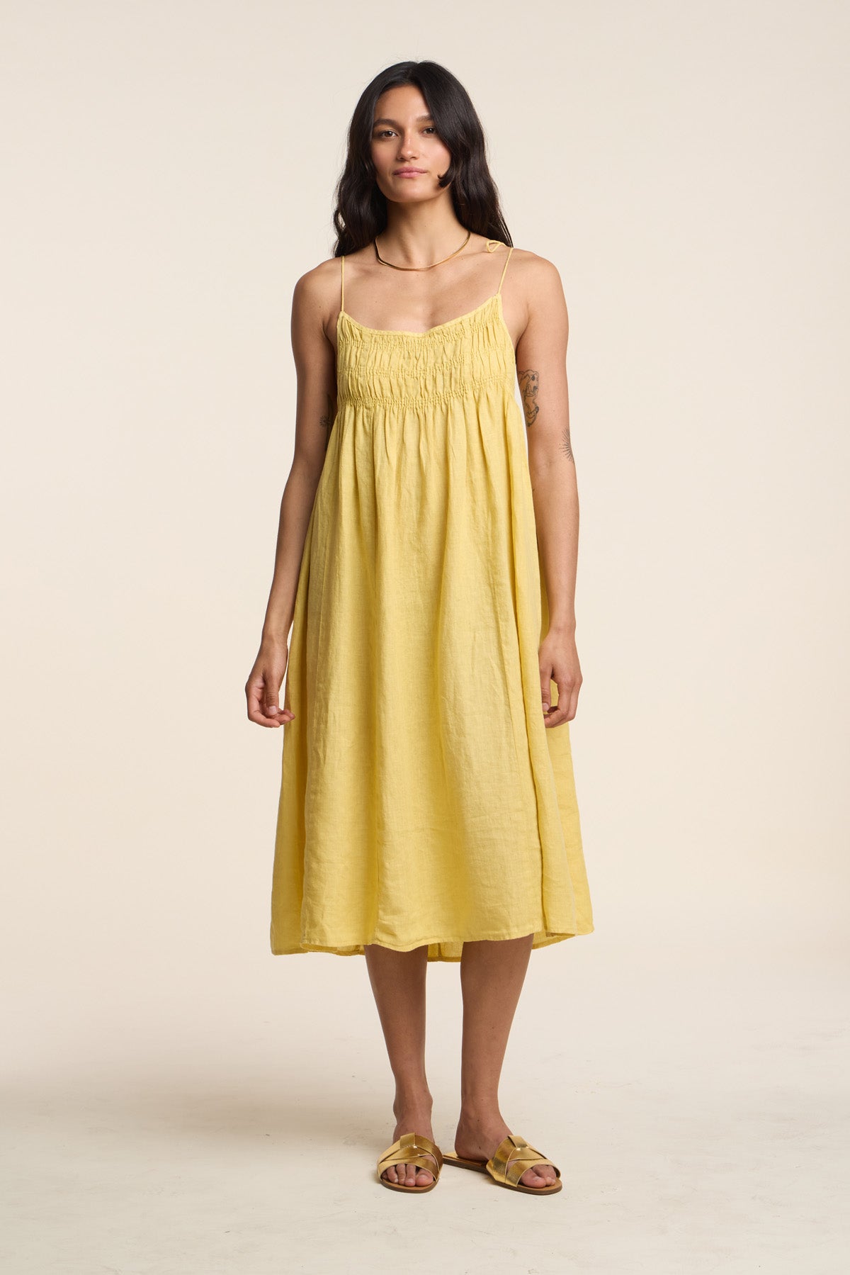 A woman wears the TERESA LINEN DRESS by Velvet by Graham & Spencer, a yellow A-line sundress with thin straps, paired with matching sandals against a plain background.