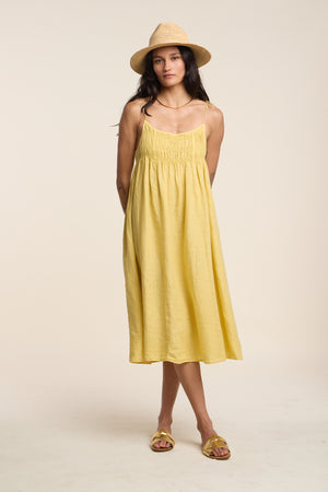 A person stands in the TERESA LINEN DRESS by Velvet by Graham & Spencer—a yellow A-line midi dress—paired with a straw hat and matching sandals against a plain light background.