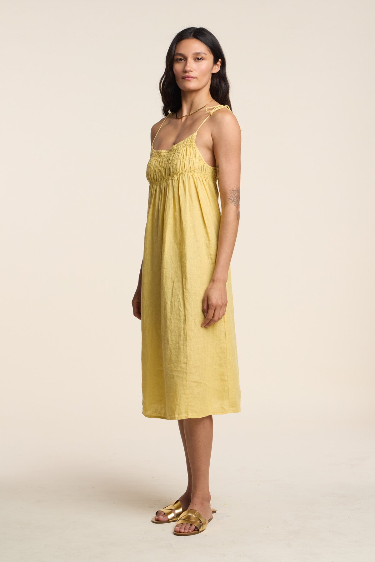 A woman stands in profile against a plain light background, wearing the TERESA LINEN DRESS by Velvet by Graham & Spencer—a yellow A-line midi dress with gathered bust—paired with gold flat sandals.