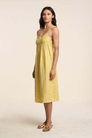 A woman stands in profile against a plain light background, wearing the TERESA LINEN DRESS by Velvet by Graham & Spencer—a yellow A-line midi dress with gathered bust—paired with gold flat sandals.