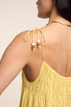 A person shown from the back and side wears the Velvet by Graham & Spencer TERESA LINEN DRESS—a yellow, sleeveless linen midi dress with beaded shoulder ties, ruched details, A-line silhouette—and a gold necklace.
