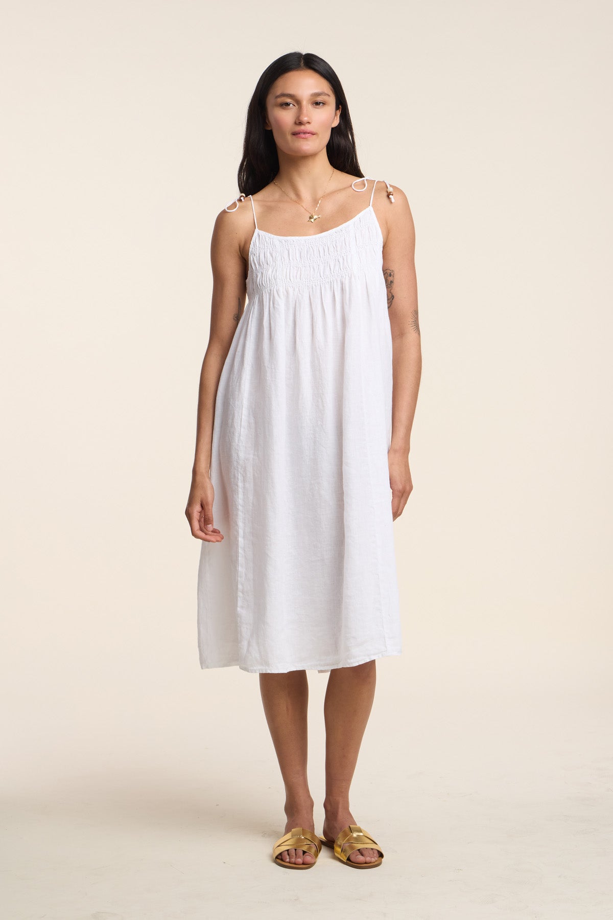 A woman faces forward in the TERESA LINEN DRESS by Velvet by Graham & Spencer, featuring smocked elastic bust and tie straps, paired with yellow sandals, set against a neutral background.-41645182648513