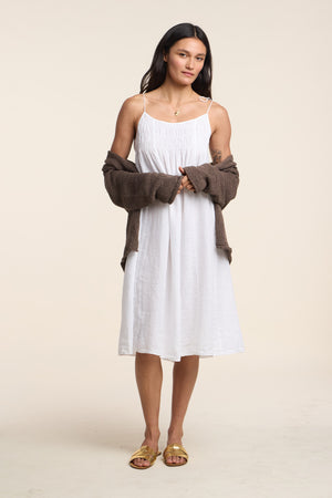A woman faces forward in the Velvet by Graham & Spencer TERESA LINEN DRESS, a white woven midi with tie straps, layered with a brown cardigan and yellow sandals, set against a neutral background.