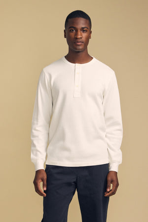 A man stands facing forward against a plain beige background, wearing the CHANCE THERMAL HENLEY by Velvet by Graham & Spencer—a white long-sleeve henley shirt—paired with dark pants.