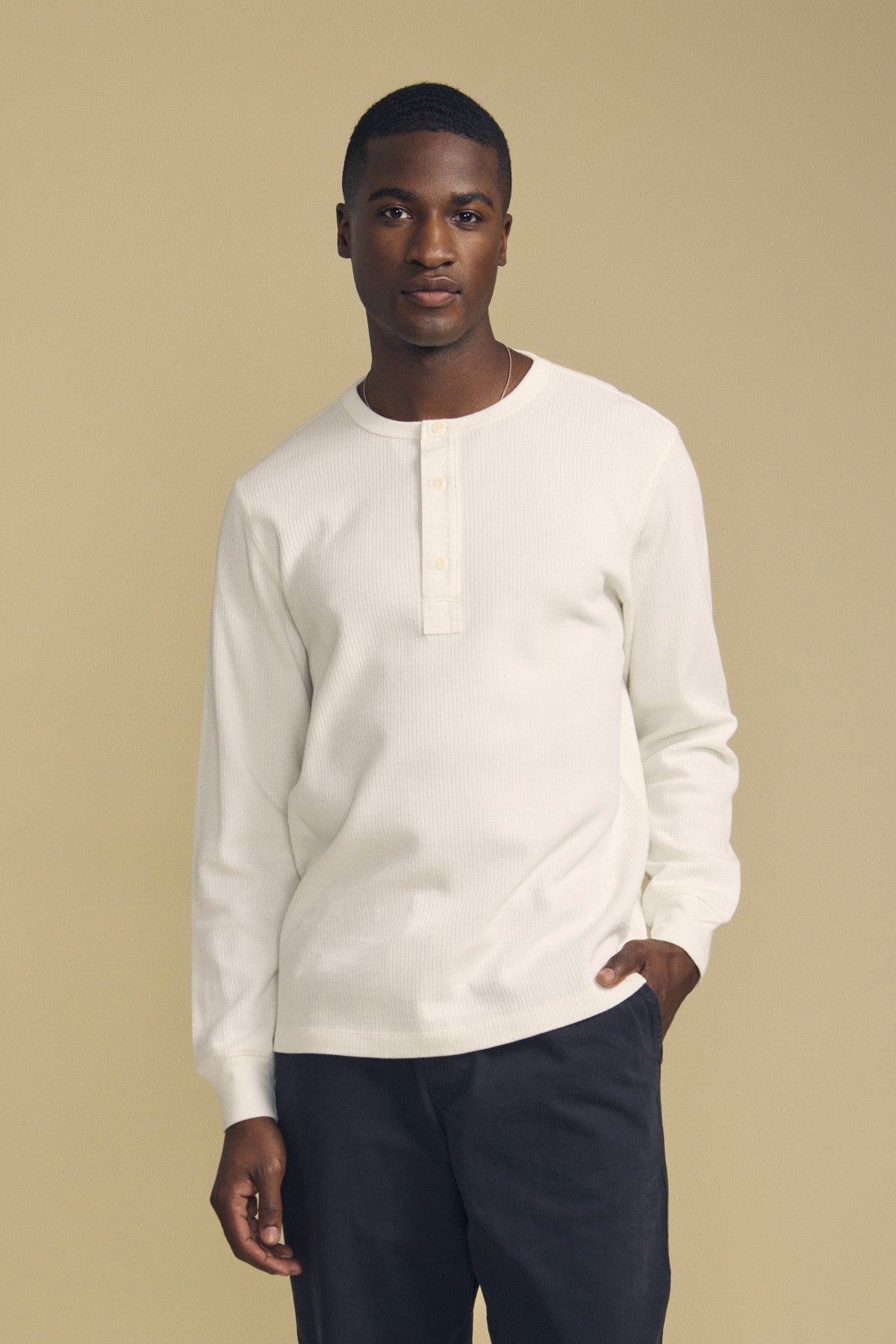 A man stands against a plain beige background, wearing the CHANCE THERMAL HENLEY by Velvet by Graham & Spencer in white and dark pants, with one hand in his pocket.-41859845128385