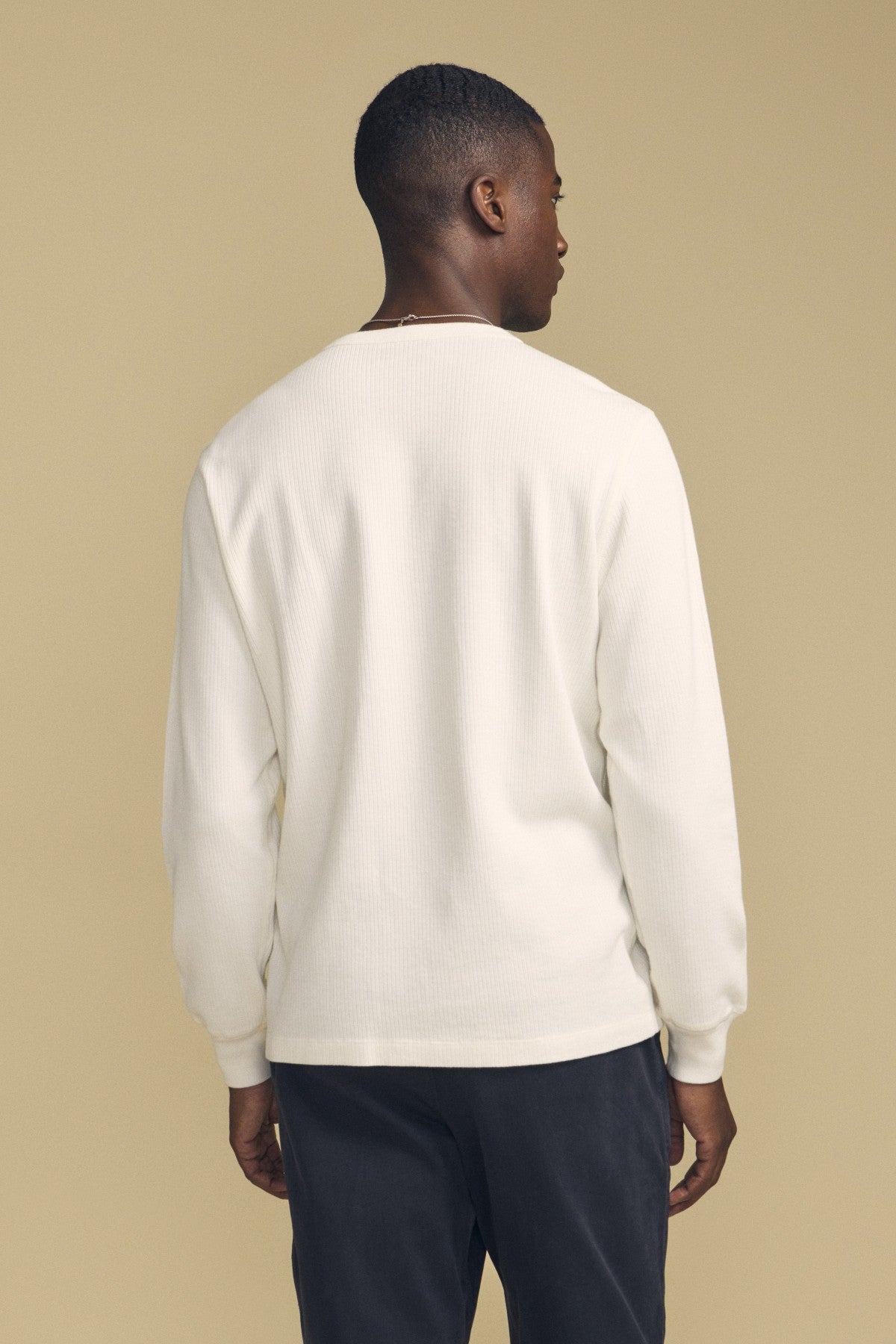 A person stands facing away, wearing the CHANCE THERMAL HENLEY by Velvet by Graham & Spencer in white with dark pants, set against a plain beige background.