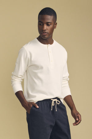 A man stands against a beige background, wearing the CHANCE THERMAL HENLEY by Velvet by Graham & Spencer and dark drawstring pants, looking slightly to the side with one hand in his pocket.