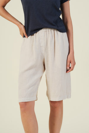 A person wears the Velvet by Graham & Spencer TOVE LINEN SHORT in light beige, featuring an elastic waist and relaxed wide-leg fit, paired with a navy short-sleeve top, standing against a plain background.