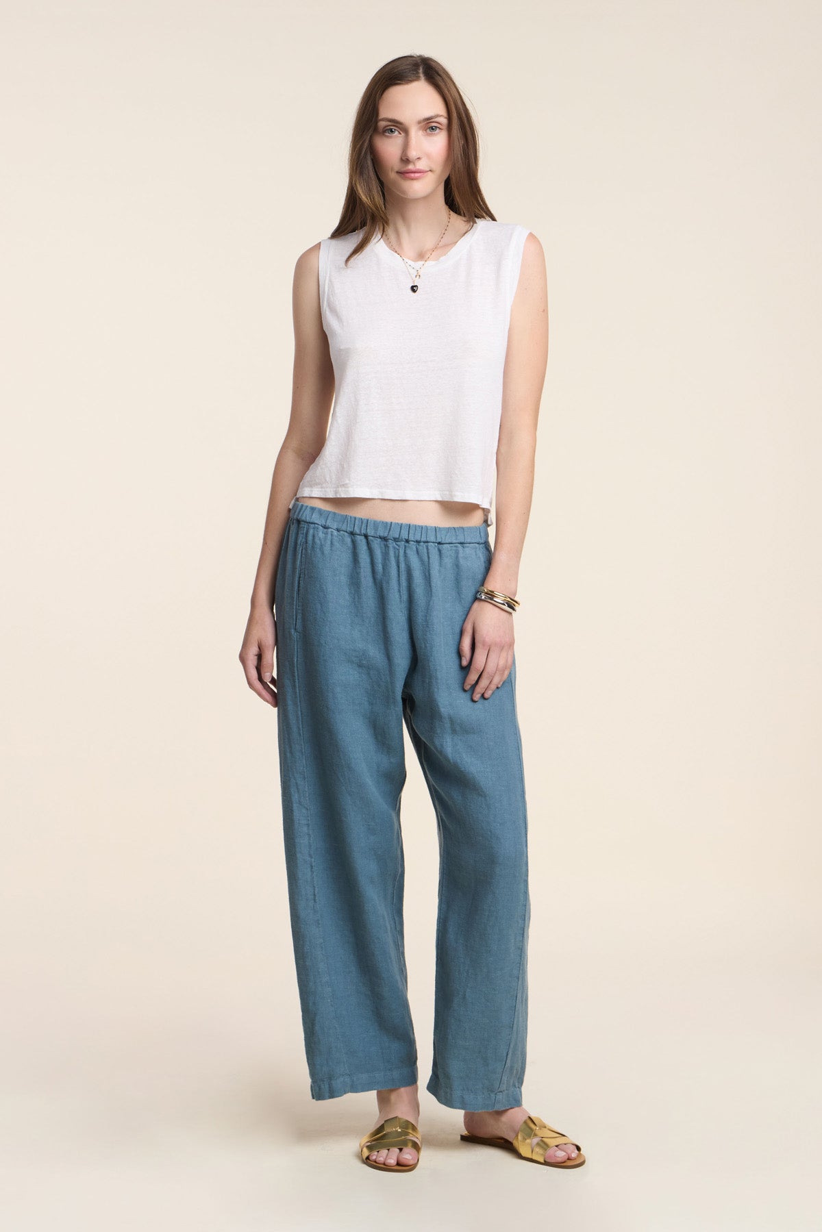 A woman faces forward against a neutral background, wearing the TRINITY TANK TOP by Velvet by Graham & Spencer—a white cropped linen knit tank with a high-low hem—paired with loose blue pants, yellow sandals, and a necklace.