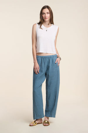 A woman stands against a plain background wearing the TRINITY TANK TOP by Velvet by Graham & Spencer as a layering piece, paired with loose blue pants, gold sandals, and a necklace.