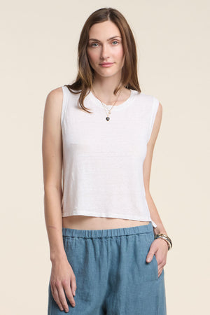 A person wears the TRINITY TANK TOP by Velvet by Graham & Spencer, a sleeveless white cropped linen tank, with blue pants, standing against a plain light background and facing the camera with a neutral expression.