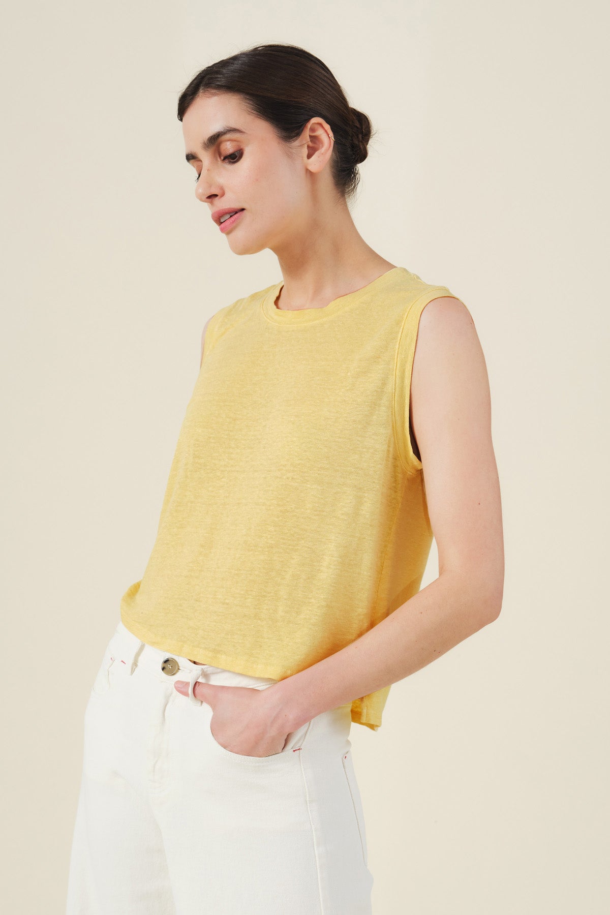 Model wearing the TRINITY TANK TOP by Velvet by Graham & Spencer in yellow, paired with white pants, standing with one hand in their pocket against a plain background.