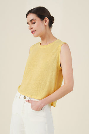 Model wearing the TRINITY TANK TOP by Velvet by Graham & Spencer in yellow, paired with white pants, standing with one hand in their pocket against a plain background.