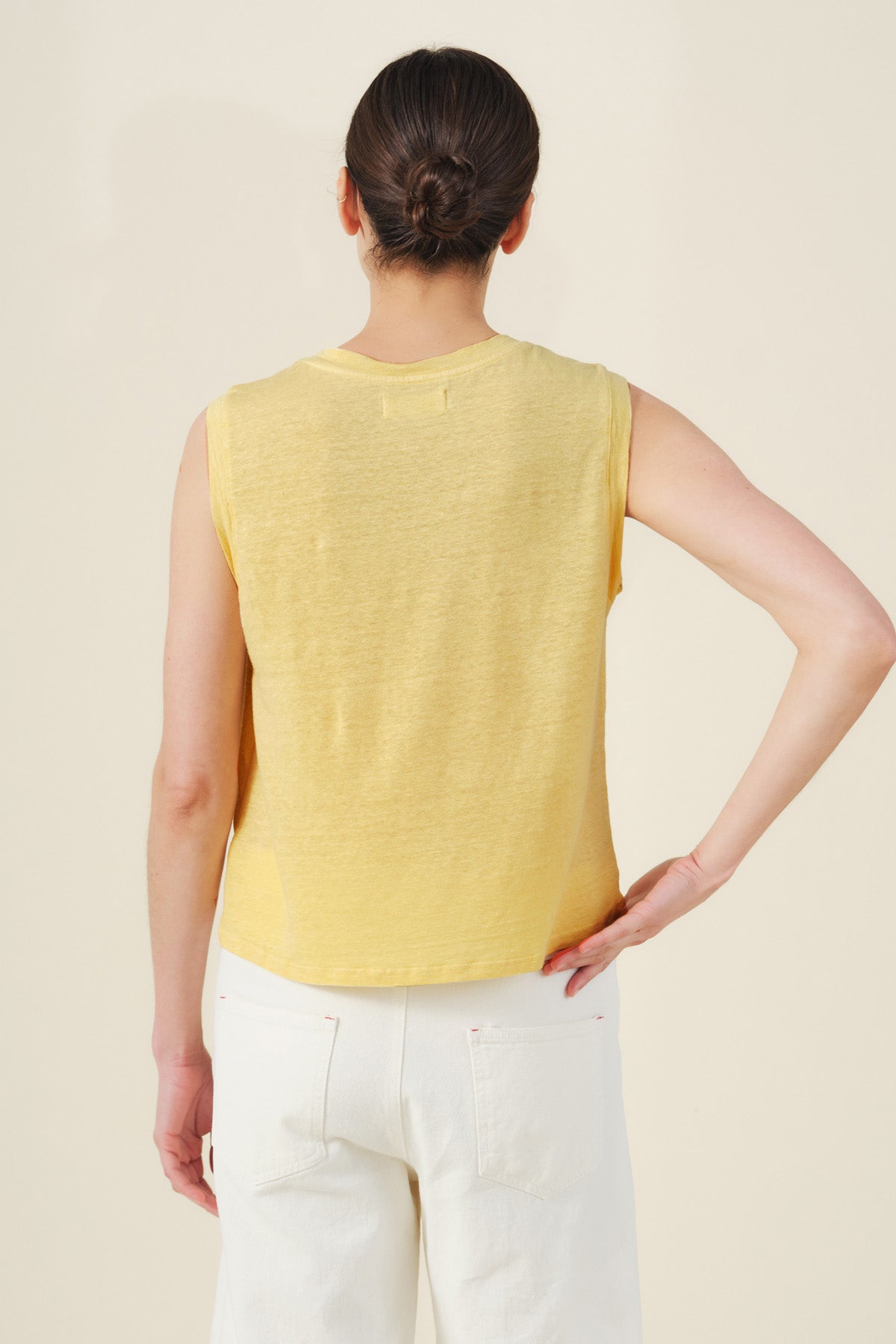 A woman with dark hair in a bun stands with her back to the camera against a plain light background, wearing the Velvet by Graham & Spencer TRINITY TANK TOP— a sleeveless, cropped linen knit tank in yellow—with white pants.