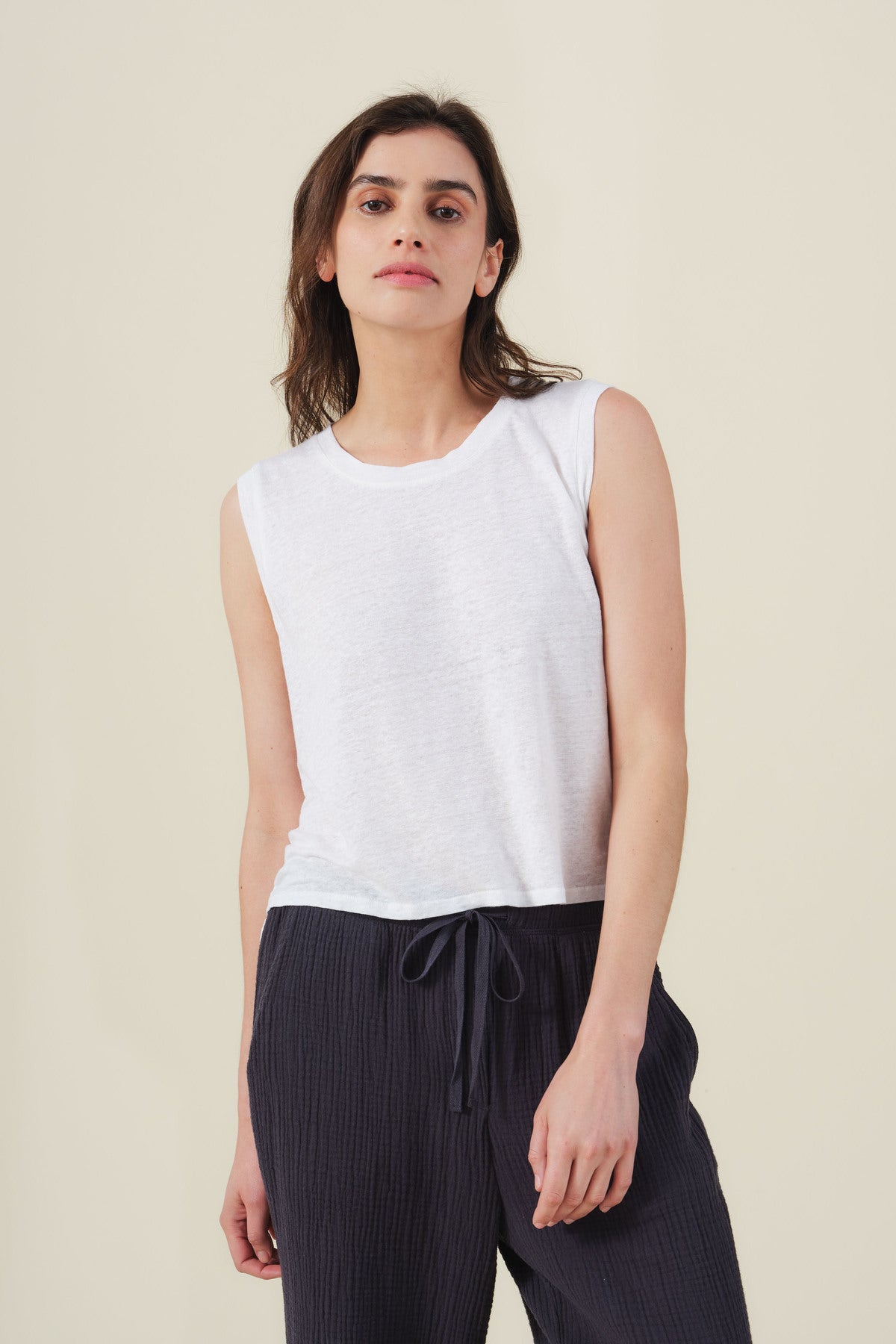A woman models the TRINITY TANK TOP by Velvet by Graham & Spencer—a cropped linen knit tank with a sheer finish—paired with dark drawstring pants, standing against a plain light background for an effortless look.