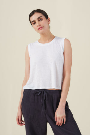 A woman wears the TRINITY TANK TOP by Velvet by Graham & Spencer—a cropped linen knit tank with a sheer finish—paired with loose dark pants, standing against a plain light background.