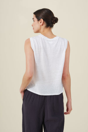 A person with hair in a low bun stands against a plain background, wearing the Velvet by Graham & Spencer TRINITY TANK TOP—a cropped linen knit tank with a sheer finish—and dark loose pants.
