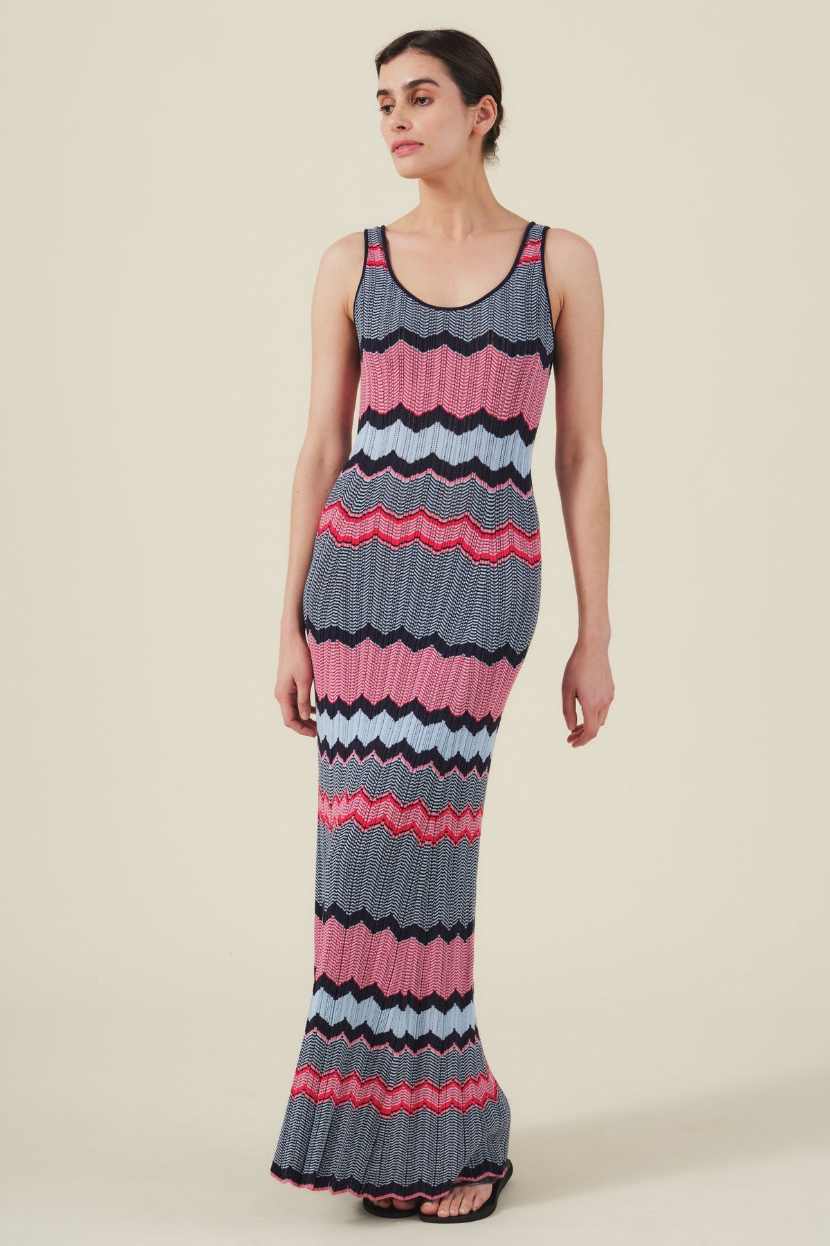 A person wears the VALENTINA DRESS by Velvet by Graham & Spencer, a sleeveless knit maxi with resort-inspired wavy stripes in blue, pink, red, black, and white, standing against a plain light background.
