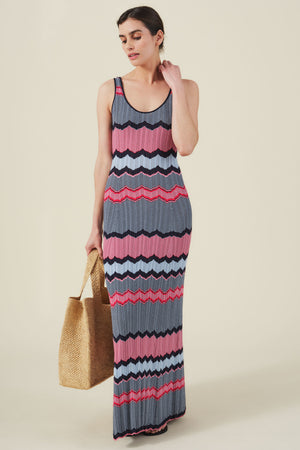A woman stands against a plain light background wearing the VALENTINA DRESS by Velvet by Graham & Spencer, a sleeveless knit maxi dress with multicolored zigzag stripes, and holds a woven tote bag.