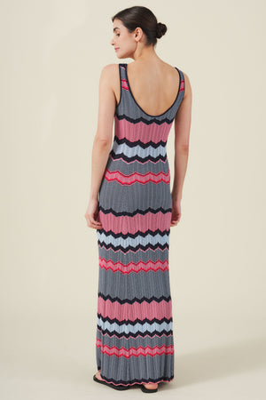 A woman stands facing away, wearing the VALENTINA DRESS by Velvet by Graham & Spencer—a resort-inspired knit maxi with wavy zigzag stripes in pink, red, blue, black, and gray.