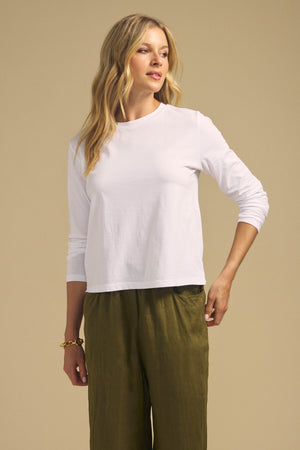 A woman wearing the VICKY TEE, a white long sleeve cotton jersey by Velvet by Graham & Spencer, and olive green pants stands against a beige background, looking to the side.