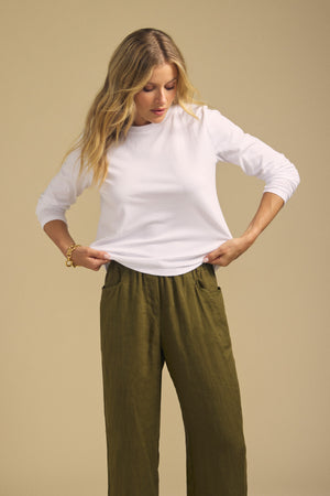 A person wearing the VICKY TEE by Velvet by Graham & Spencer, paired with olive green pants, stands against a beige background, looking down while holding the bottom of their shirt.