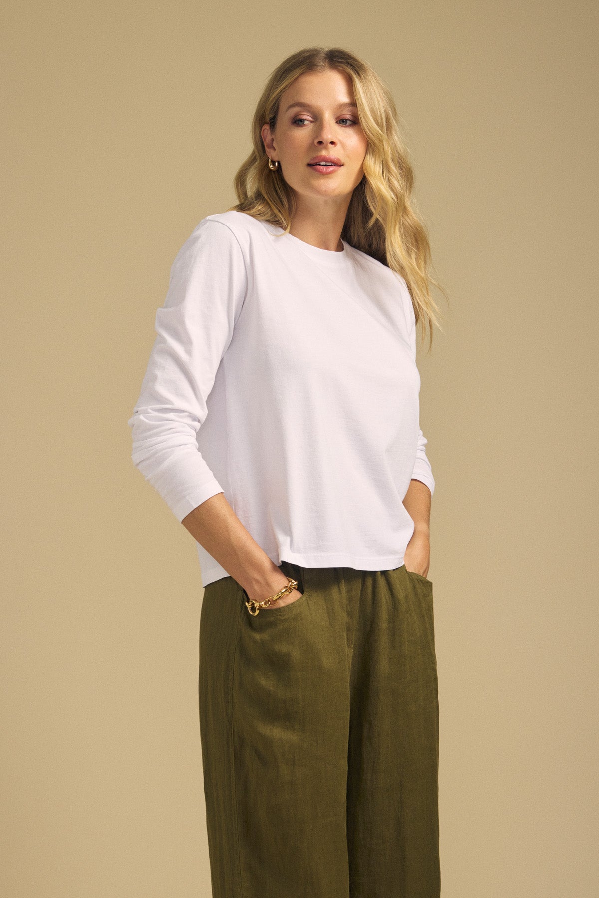 A woman stands against a beige background, wearing the VICKY TEE by Velvet by Graham & Spencer— a white long sleeve cotton jersey tee—paired with olive green wide-leg pants, her hands in her pockets.