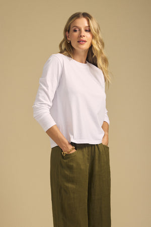 A woman stands against a beige background, wearing the VICKY TEE by Velvet by Graham & Spencer— a white long sleeve cotton jersey tee—paired with olive green wide-leg pants, her hands in her pockets.