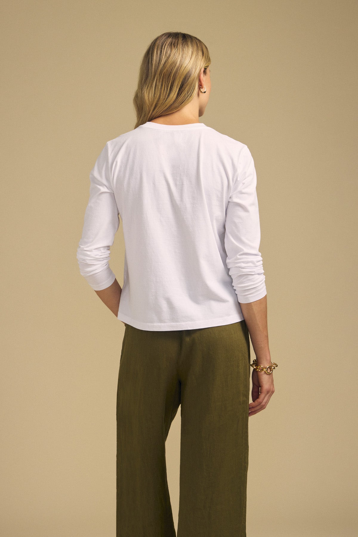 A woman stands facing away against a plain beige background, wearing the VICKY TEE—a long sleeve white cotton jersey tee by Velvet by Graham & Spencer—and olive green pants.-41929472540865