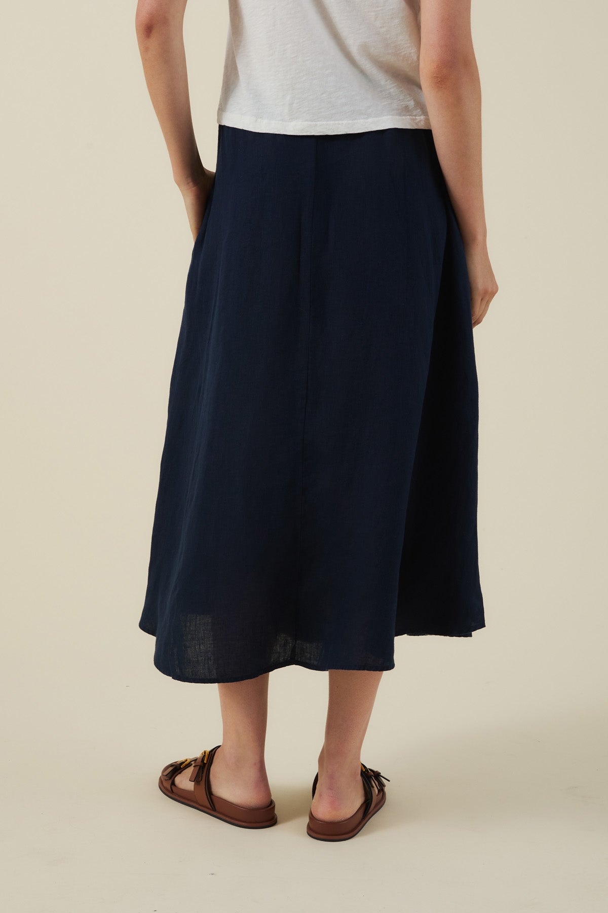 Model shown from the back wearing a white short-sleeve top, brown sandals, and the VIRGINIA LINEN SKIRT by Velvet by Graham & Spencer—an A-line navy blue woven linen skirt with an elastic waist—against a light plain background.