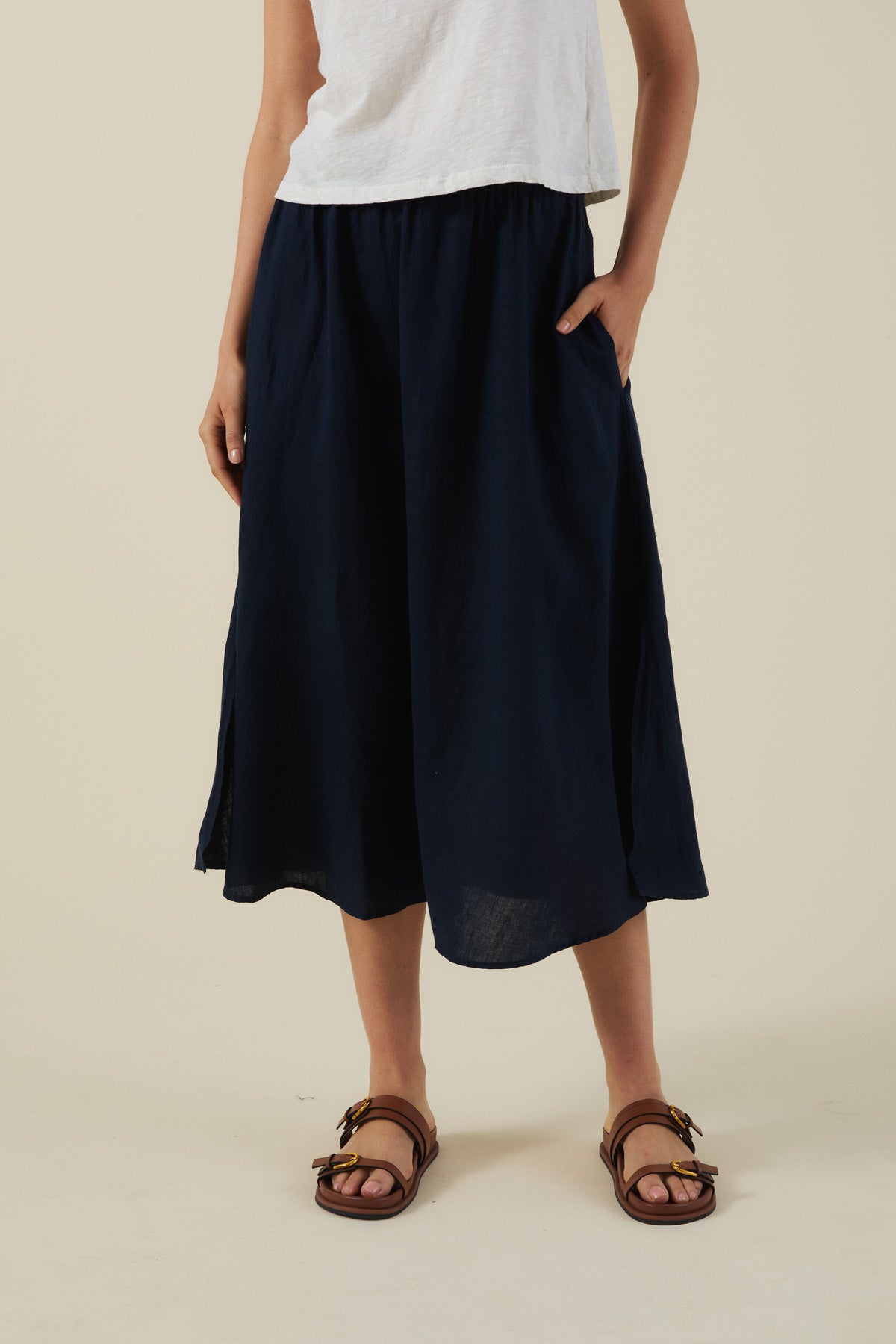 A person wears a white t-shirt, brown sandals, and the VIRGINIA LINEN SKIRT by Velvet by Graham & Spencer—an A-line navy blue woven linen midi skirt with elastic waist and pockets—standing against a plain, light-colored background.