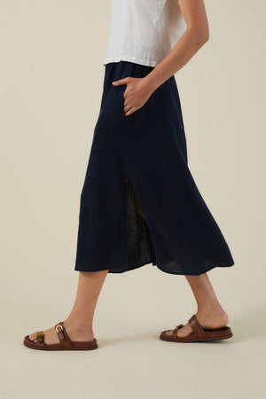A person walks against a plain beige background in a white top, brown sandals, and the VIRGINIA LINEN SKIRT by Velvet by Graham & Spencer—an A-line navy blue midi skirt with a side slit.
