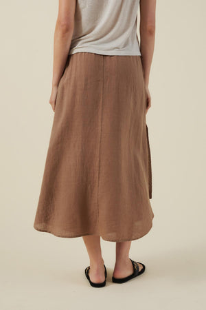 A person wearing a light gray T-shirt, black sandals, and the VIRGINIA LINEN SKIRT by Velvet by Graham & Spencer stands with their back to the camera against a plain background.