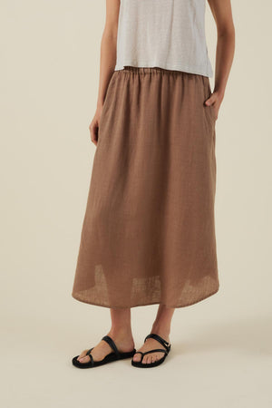 A person wearing a white sleeveless top, the VIRGINIA LINEN SKIRT by Velvet by Graham & Spencer—featuring brown woven linen, an A-line midi cut, pockets, and an elastic waist—and black sandals stands against a plain light background.