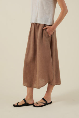 A person wears a white sleeveless top, black sandals, and the VIRGINIA LINEN SKIRT by Velvet by Graham & Spencer—an A-line light brown linen midi skirt with pockets and elastic waist—standing against a plain background.