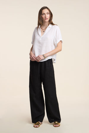 A woman faces forward wearing the VIVICA LINEN TOP by Velvet by Graham & Spencer, paired with wide black pants, gold sandals, and minimal jewelry—an easy, stylish look for warm days against a simple light background.
