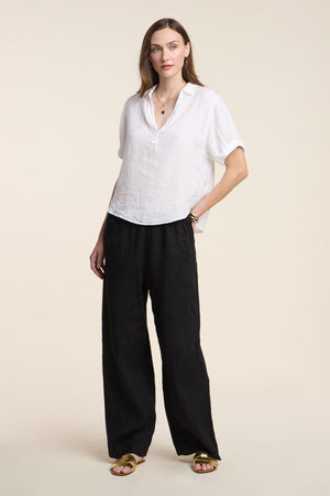 A woman wears the VIVICA LINEN TOP by Velvet by Graham & Spencer, styled with loose black pants and gold sandals—an easy, breezy look for warm weather set against a simple light backdrop.
