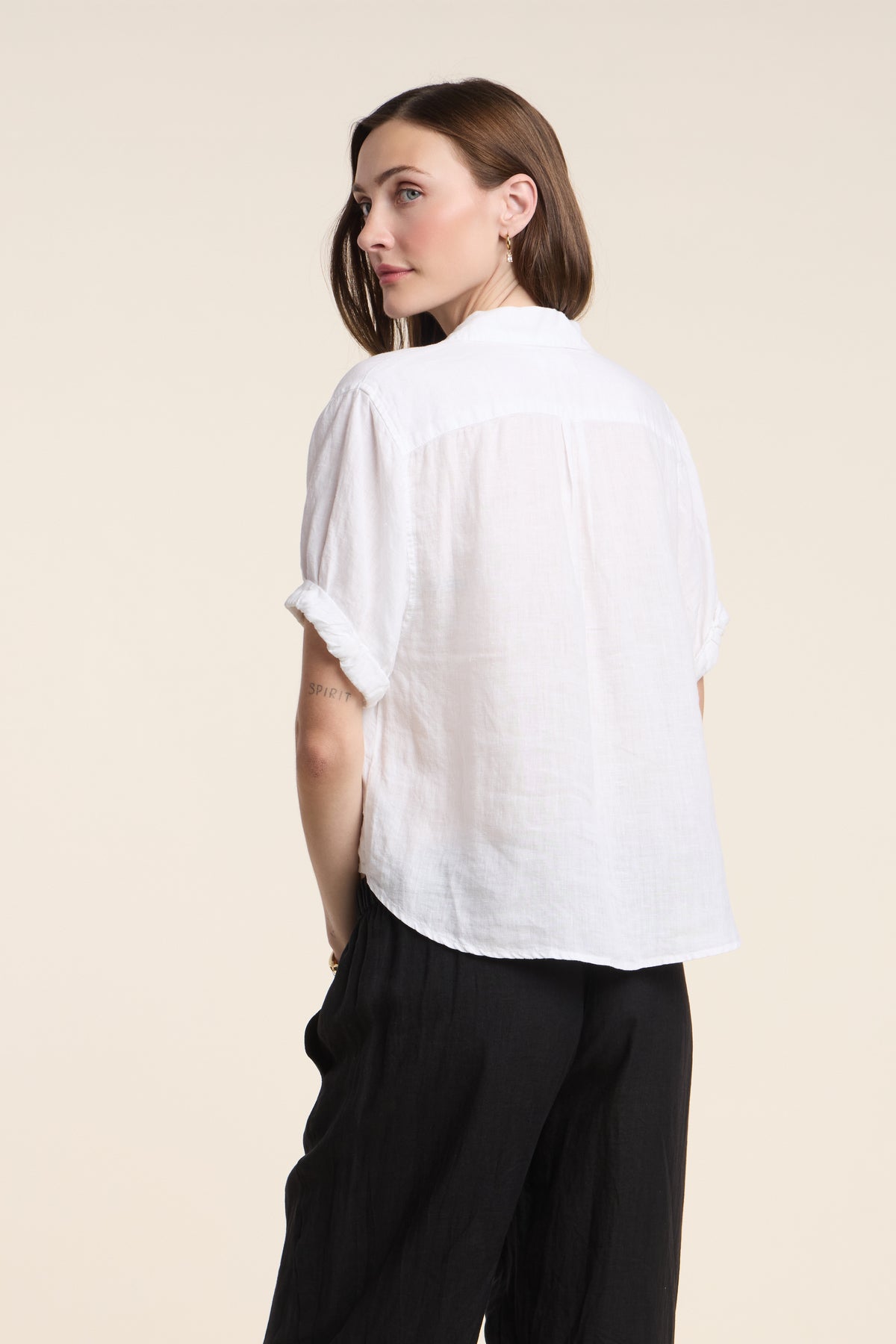 A woman stands with her back to the camera in the VIVICA LINEN TOP by Velvet by Graham & Spencer—an airy short-sleeve linen piece styled with black pants, ideal for warm weather, set against a neutral background.
