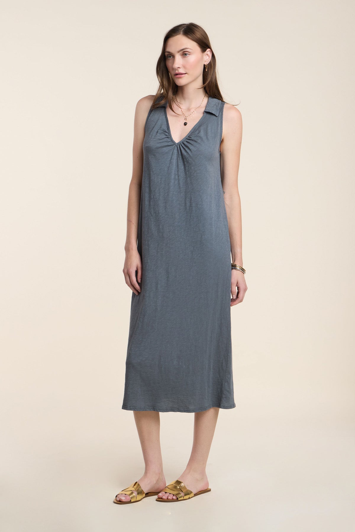 A woman stands in a studio against a beige background, wearing the YORK DRESS by Velvet by Graham & Spencer—a sleeveless, knee-length grey cotton slub dress with a V-neckline—paired with gold sandals.