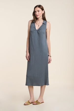 A woman stands in a studio against a beige background, wearing the YORK DRESS by Velvet by Graham & Spencer—a sleeveless, knee-length grey cotton slub dress with a V-neckline—paired with gold sandals.