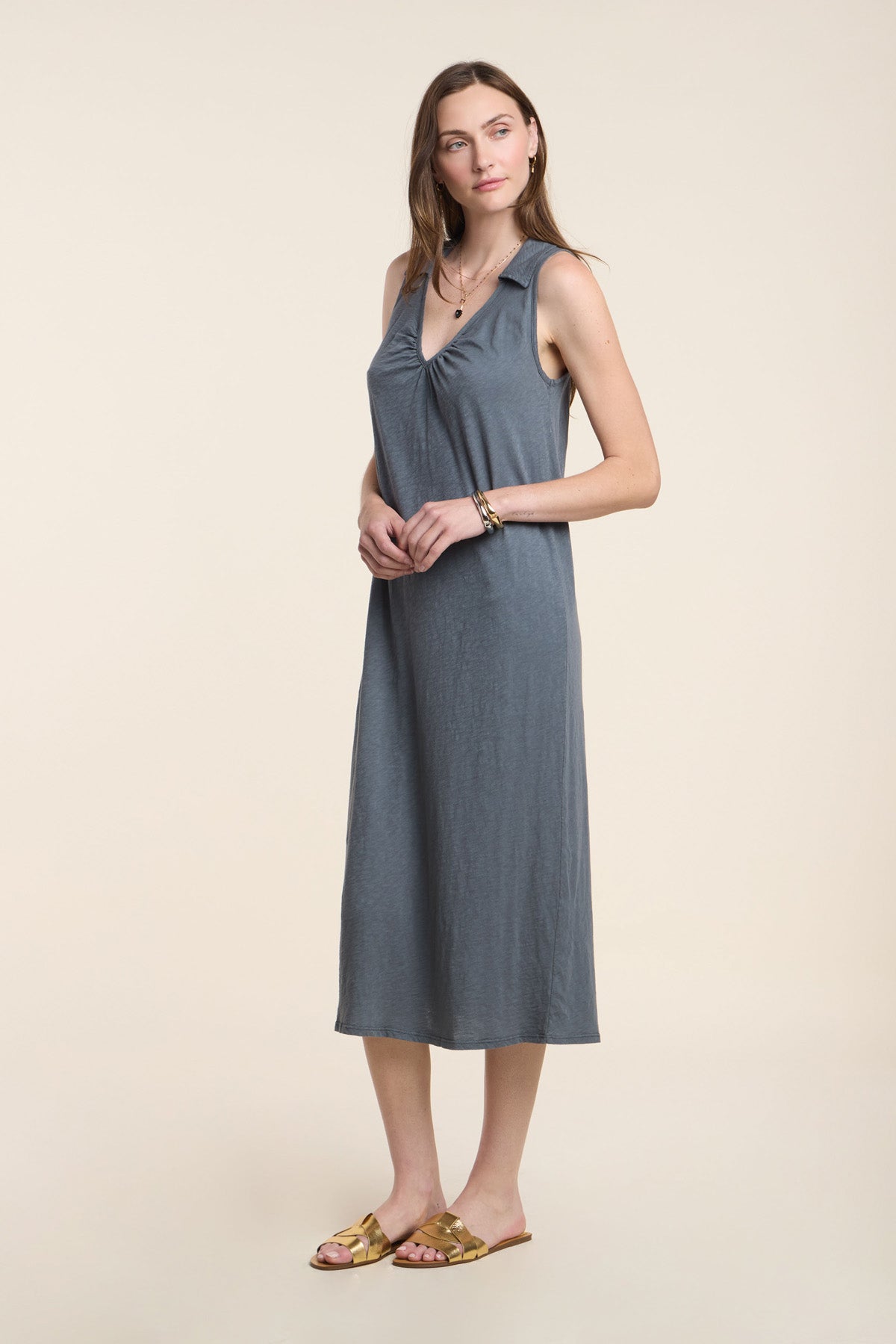 A woman stands against a plain background, wearing the YORK DRESS by Velvet by Graham & Spencer—a sleeveless gray cotton slub midi dress—paired with flat sandals, minimal jewelry, and natural makeup.