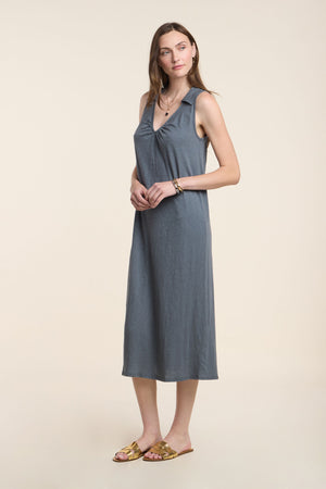 A woman stands against a plain background, wearing the YORK DRESS by Velvet by Graham & Spencer—a sleeveless gray cotton slub midi dress—paired with flat sandals, minimal jewelry, and natural makeup.