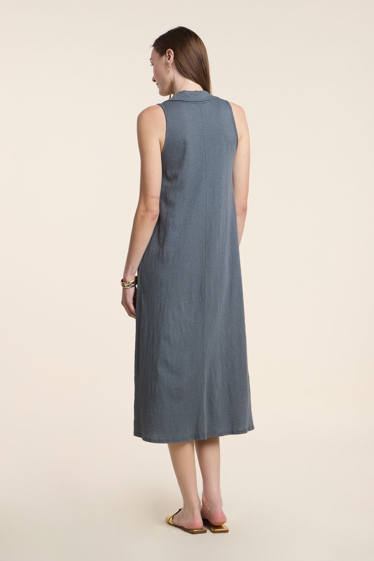 A woman stands with her back to the camera, wearing the YORK DRESS by Velvet by Graham & Spencer—a sleeveless gray midi dress—paired with flat sandals against a plain, light background.