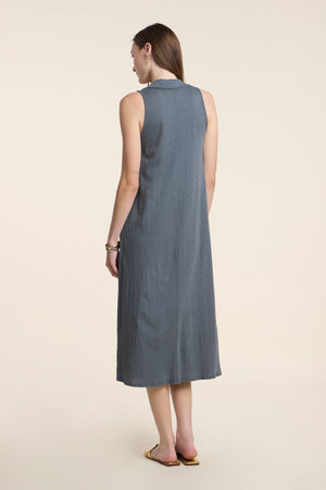 A woman stands with her back to the camera, wearing the YORK DRESS by Velvet by Graham & Spencer—a sleeveless gray midi dress—paired with flat sandals against a plain, light background.